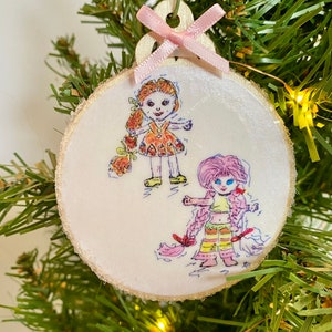 May include: A white wooden ornament with a pink ribbon and a hand-drawn illustration of two girls with long hair. The girls are wearing colorful dresses and are standing on a white background.
