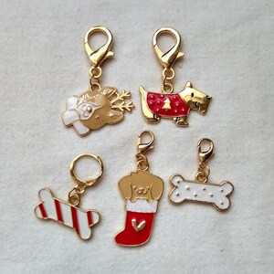 May include: Five gold-tone metal Christmas-themed charms with clip-on attachments. The charms feature a reindeer, a dog wearing a red sweater, a dog in a stocking, a candy cane bone, and a plain bone.