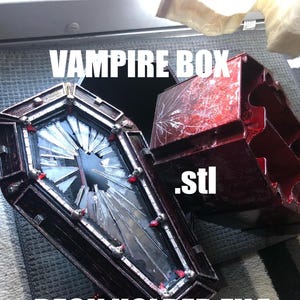 May include: A red and black coffin-shaped box with a broken glass window. The box is labeled "Vampire Box .stl Deck Holder File".