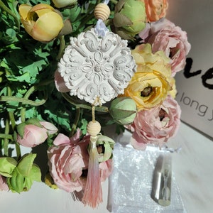May include: White ceramic ornament with a floral design, hanging from a white string with a wooden bead and a pink tassel. The ornament is surrounded by pink and yellow flowers.