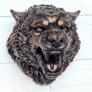 Könnte beinhalten: Eine bronzefarbene Wolfskopfskulptur mit offenem Maul, das scharfe Zähne zeigt. Die Skulptur hat detailliertes Fell und intensive Augen, montiert an einer weißen Ziegelmauer. Dieses dekorative Stück ist ein Beispiel für Tierkunst.