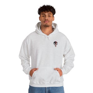 May include: A light gray hoodie with a front pocket and a hood. The hoodie features a small embroidered skull design on the upper left chest, with black, red, and white details. The model is wearing blue jeans.