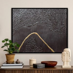 May include: Abstract painting with a textured black background and a gold line forming a mountain shape. The painting is framed in black and displayed on a shelf with other decorative items.