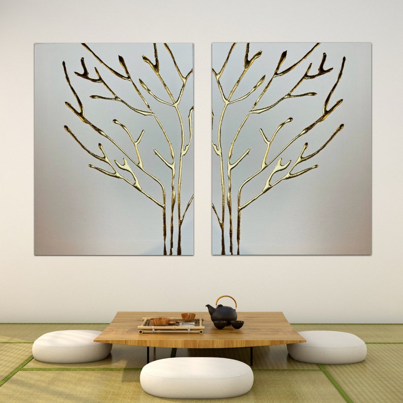May include: Two gold leaf paintings of stylized trees on a white wall above a wooden table with two white cushions and a black teapot with a lid.