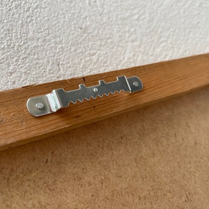 May include: A close-up of a wooden picture frame with a metal hanging hook attached to the back.