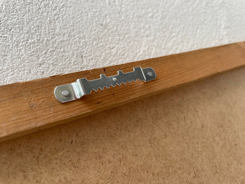 May include: A close-up of a wooden picture frame with a metal hanging hook attached to the back.