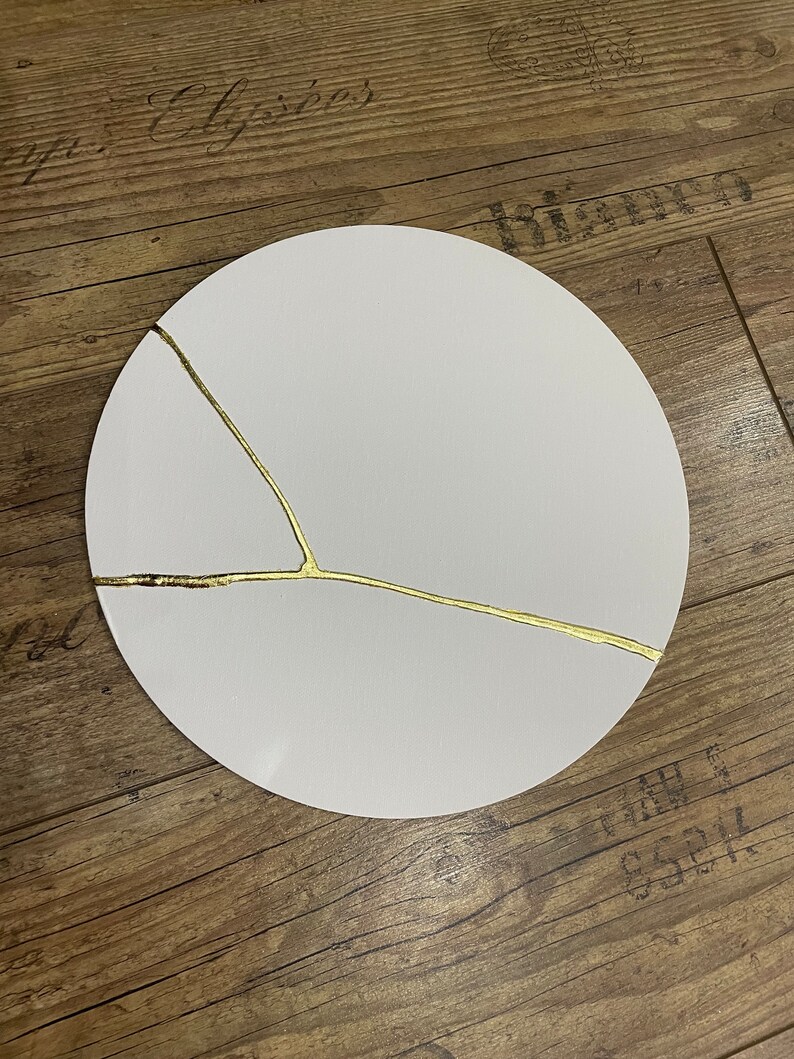 Puede incluir: Un plato redondo blanco con un dise&ntilde;o de craquelado dorado. El plato est&aacute; sobre una superficie de madera.