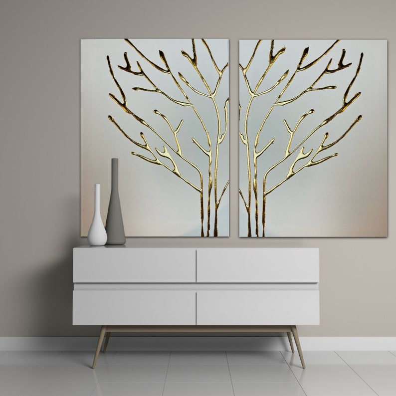 May include: Two white canvases with gold leaf tree branches on a light gray background. The canvases are hanging above a white dresser with four drawers and wooden legs.