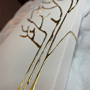 May include: A white canvas with a gold tree design painted on the front. The canvas is wrapped around the edges and has a white background.