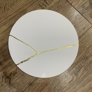 Puede incluir: Un plato redondo blanco con un dise&ntilde;o de craquelado dorado. El plato est&aacute; sobre una superficie de madera.