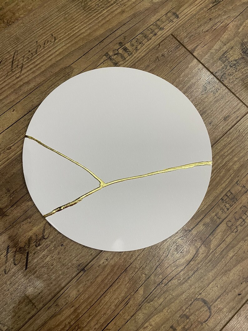 Puede incluir: Un plato redondo blanco con un dise&ntilde;o de craquelado dorado. El plato est&aacute; sobre una superficie de madera.