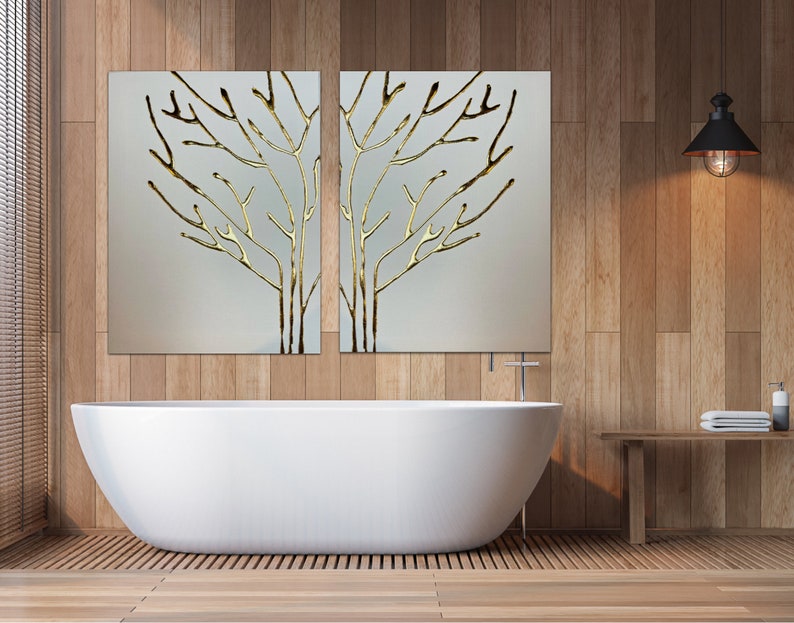 May include: A white bathtub sits in a bathroom with wood paneling on the walls. Two framed pieces of art with gold leaf tree branches hang above the tub. A wooden shelf with a towel and a bottle of soap sits on the right side of the image.