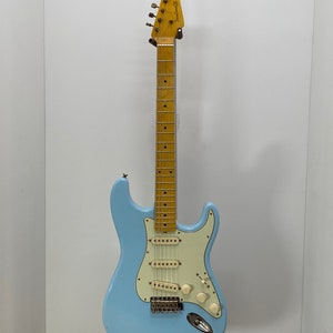 Könnte beinhalten: Eine hellblaue E-Gitarre mit weißem Schlagbrett und einem Ahornhals. Die Gitarre hat ein Fender-Logo auf dem Kopf.