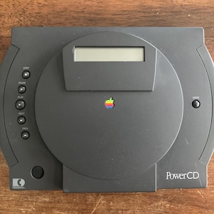 May include: A dark gray Apple PowerCD player with a circular design. It features a display screen, control buttons, and the Apple logo in the center. The text "PowerCD" is visible on the lower right.