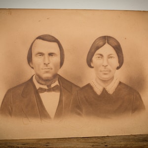 May include: A sepia-toned photograph of a couple, a man and a woman, posed formally in front of a plain background. The man is wearing a dark suit with a bow tie, and the woman is wearing a dark dress with a lace collar.