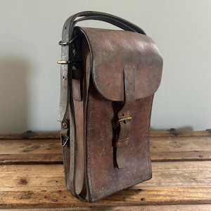 May include: A tall, rectangular brown leather bag with a weathered texture. It features a top handle, a fold-over flap secured by a single leather strap with a brass-toned metal buckle, and an adjustable side strap with brass-toned hardware.