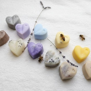 Könnte beinhalten: Eine Auswahl herzförmiger Wachsschmelzen in verschiedenen Farben wie Grau, Lavendel, Blau und Gelb, auf einer weißen Oberfläche angeordnet. Ein Lavendelzweig und zwei Bienen sind ebenfalls zu sehen. Die Schmelzen haben ein rustikales Aussehen.
