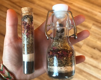 May include: Two glass bottles filled with dried herbs, flowers, and crystals. The smaller bottle has a cork stopper, while the larger bottle has a metal clasp.