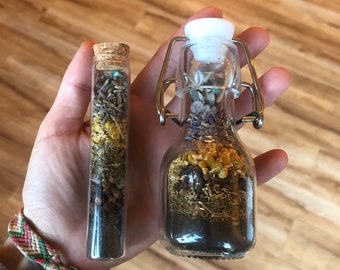 May include: Two glass bottles filled with dried herbs and flowers. The smaller bottle has a cork stopper and the larger bottle has a metal clasp.