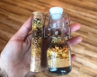 May include: Two glass bottles filled with dried herbs and flowers. The smaller bottle has a cork stopper and the larger bottle has a metal wire closure.