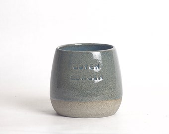 Könnte beinhalten: Handgefertigte Keramiktasse mit zweifarbigem Design. Die obere Hälfte ist ein gedämpftes Blau-Grau, die untere Hälfte hellbeige. Auf der Vorderseite steht "Guten Morgen". Die Tasse hat eine abgerundete Form und eine glatte Oberfläche.
