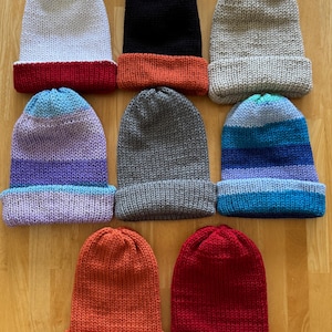 May include: A collection of nine knitted beanies in various colors, including white with a red brim, black with an orange brim, and gray. Other beanies feature blue, purple, and orange color schemes. The beanies are displayed on a wooden surface.