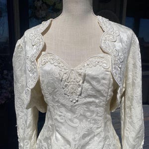 May include: Ivory-colored wedding dress with a matching bolero jacket. The dress features a sweetheart neckline and intricate floral embroidery. The bolero has long sleeves and delicate lace trim, creating a vintage bridal look.