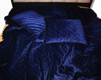 May include: A navy blue velvet bedspread with two matching pillows. The bedspread and pillows have a quilted pattern. The pillows are trimmed with a dark blue pom-pom fringe. The bed is covered with a luxurious, soft, and inviting bedding set.