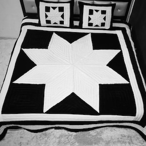Pode incluir: Conjunto de edredão a preto e branco com um padrão de estrela. O edredão apresenta uma grande estrela branca no centro, rodeada por uma borda preta. Duas almofadas quadradas a condizer com motivos de estrelas completam o conjunto. O conjunto está numa cama com cabeceira preta.