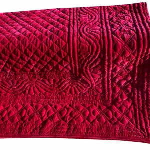 Op de afbeelding: Een dieprode fluwelen quilt met ingewikkelde gewatteerde patronen. Het ontwerp heeft ruit-, bloemen- en geometrische vormen. De pluche stof is opgevouwen en toont de gedetailleerde stiksels en textuur van het beddengoed.