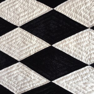 Puede incluir: Primer plano de un textil acolchado con un patrón de diamantes repetido. El diseño alterna entre diamantes negros y blancos, creando un efecto visual de alto contraste. Los diamantes blancos tienen una apariencia texturizada y acolchada.