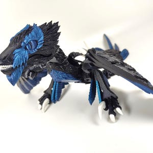 May include: A detailed figurine of a dragon with a black body and blue accents. The dragon has a feathered head, wings, and tail. Its claws and teeth are white. The figurine is posed on a white surface.