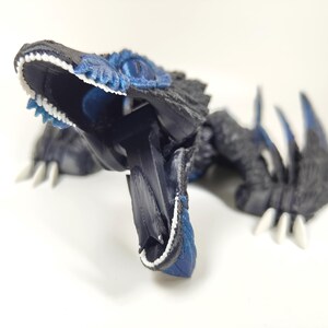 May include: A detailed, 3D-printed dinosaur figurine in shades of black and blue, with white teeth and claws. The open-mouthed creature has textured details and a dynamic pose, showcasing its sharp teeth and claws.