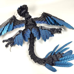 May include: A detailed, articulated figurine of a mythical creature with a bird-like form. The figure features a black body with blue accents on the wings, tail, and other feathered areas. The wings are spread, and the figure is posed on a white surface.