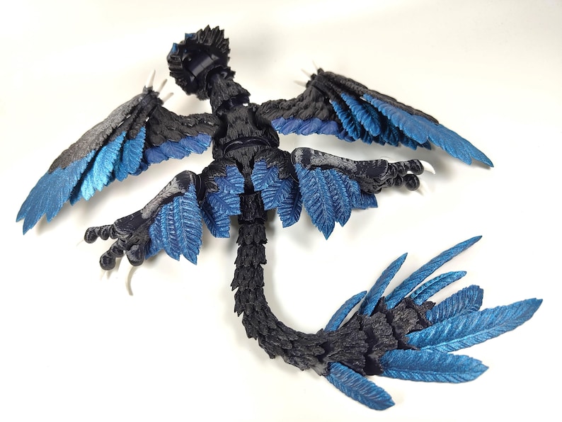 May include: A detailed, articulated figurine of a mythical creature with a bird-like form. The figure features a black body with blue accents on the wings, tail, and other feathered areas. The wings are spread, and the figure is posed on a white surface.