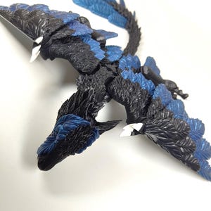 May include: A detailed sculpture of a mythical creature with a dragon-like form. The figure features black and blue feathered wings and body, with white accents on the beak-like head. The sculpture is positioned on a white surface.