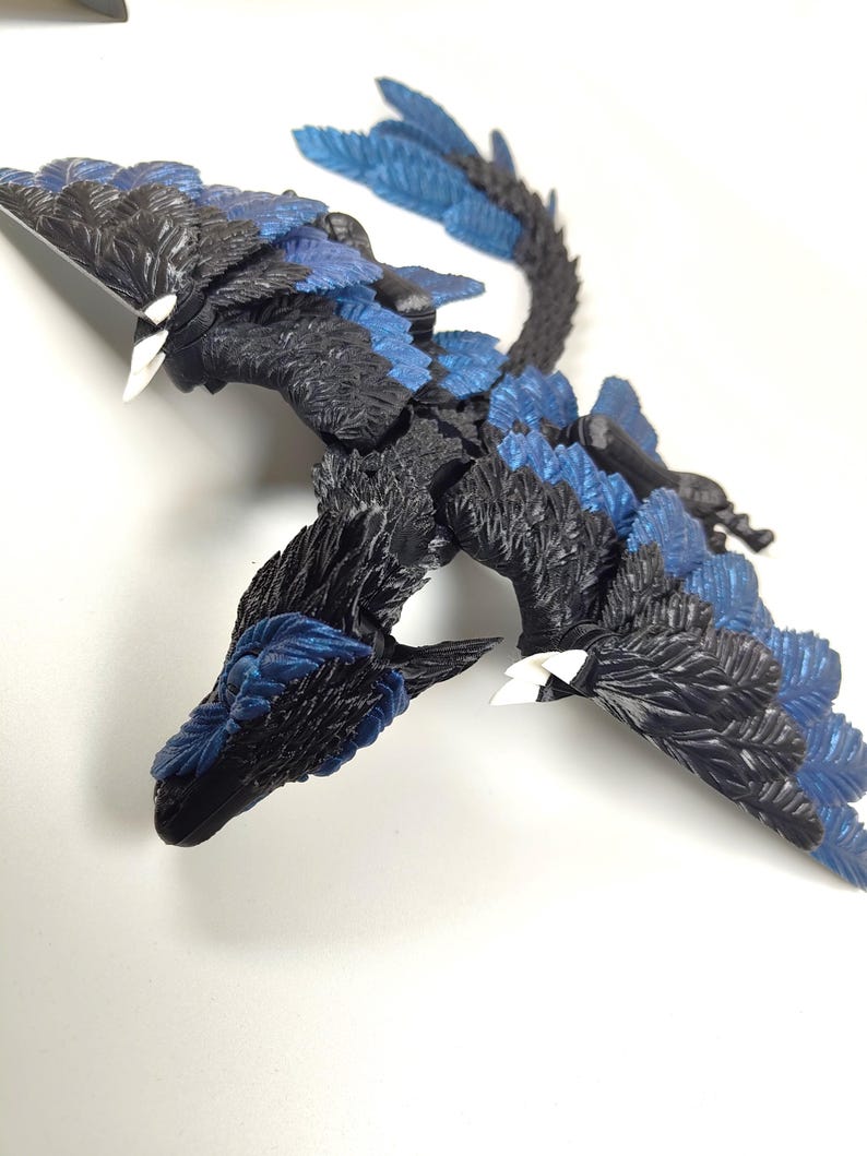 May include: A detailed sculpture of a mythical creature with a dragon-like form. The figure features black and blue feathered wings and body, with white accents on the beak-like head. The sculpture is positioned on a white surface.