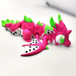 Puede incluir: Una figurita de dragón rosa y verde con manchas blancas, que se asemeja a un fruto del dragón. El dragón está acostado de lado con las alas extendidas.