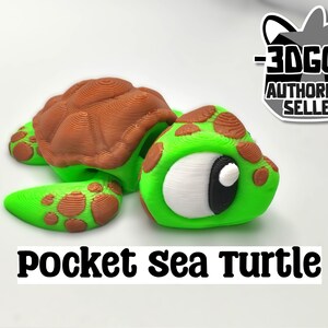 Puede incluir: Una pequeña figura de tortuga marina verde y marrón con un ojo blanco y una pupila negra. La tortuga está tumbada boca abajo con la cabeza girada hacia un lado. El texto "Pocket Sea Turtle" está impreso debajo de la tortuga.