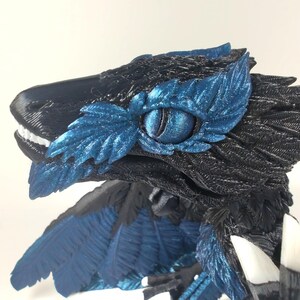 May include: A detailed, 3D-printed sculpture of a raptor-like creature. The sculpture features a black head and body with blue accents around the eye and on the wings. The creature has white teeth and a detailed eye.