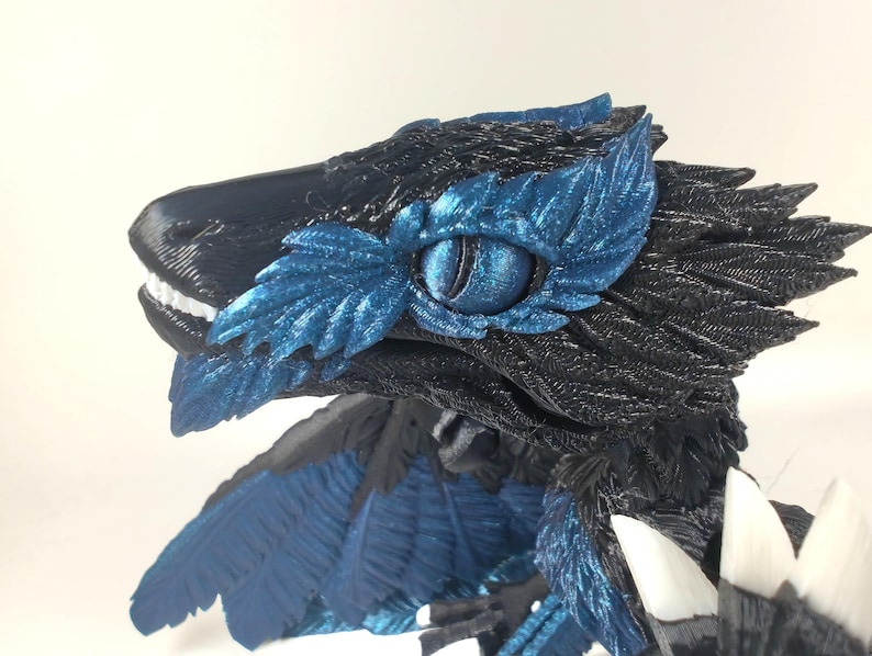 May include: A detailed, 3D-printed sculpture of a raptor-like creature. The sculpture features a black head and body with blue accents around the eye and on the wings. The creature has white teeth and a detailed eye.