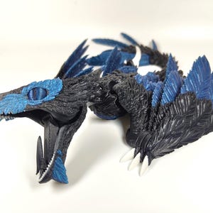 May include: A detailed, 3D-printed dragon figurine in shades of black and blue. The dragon features textured scales, feathered wings, and sharp white claws. Its head is open, revealing teeth and a detailed interior. The eyes are a striking blue.