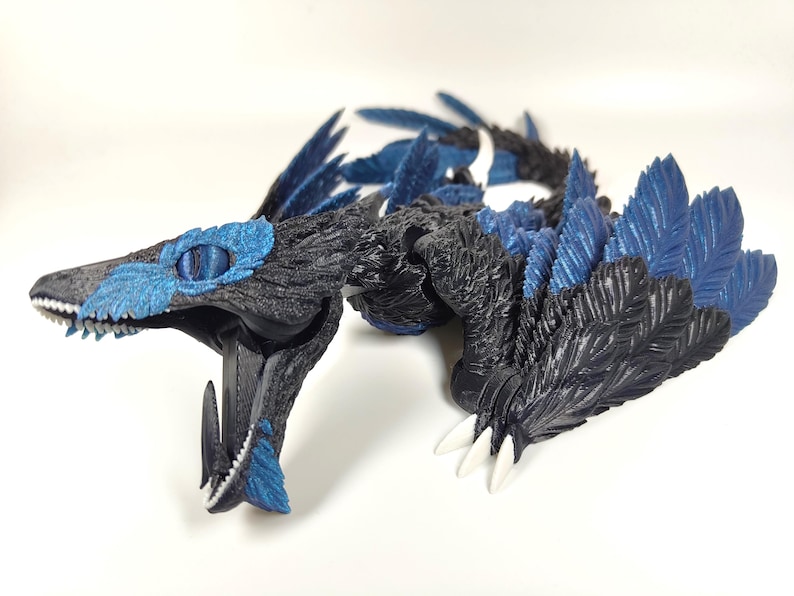 May include: A detailed, 3D-printed dragon figurine in shades of black and blue. The dragon features textured scales, feathered wings, and sharp white claws. Its head is open, revealing teeth and a detailed interior. The eyes are a striking blue.