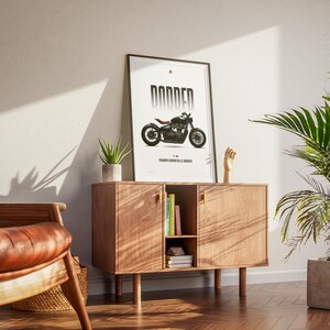 May include: A framed poster with a motorcycle illustration, titled "DODDER," rests on a wooden cabinet. The cabinet has two doors and a central open shelf with books. A potted plant and a wooden hand sculpture are also on the cabinet.