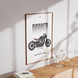 May include: Framed poster featuring a black motorcycle with the word "BONNER" in bold, vertical letters. The motorcycle is a Triumph Bonneville Bobber. The poster has a wooden frame and is displayed on a white wall.