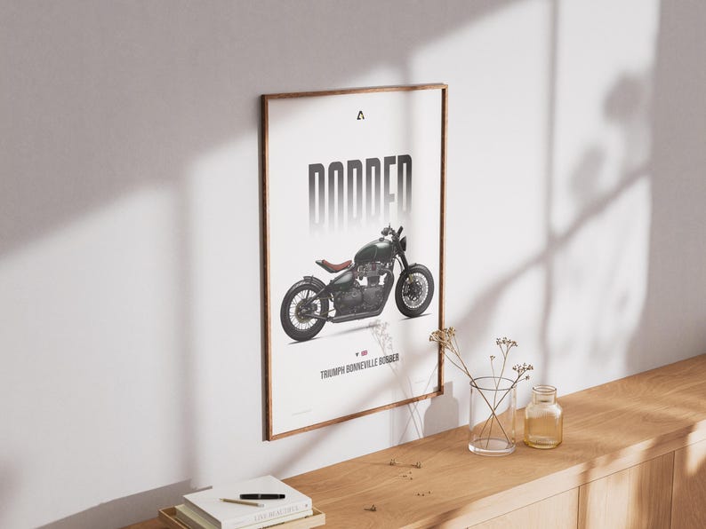 May include: Framed poster featuring a black motorcycle with the word "BONNER" in bold, vertical letters. The motorcycle is a Triumph Bonneville Bobber. The poster has a wooden frame and is displayed on a white wall.