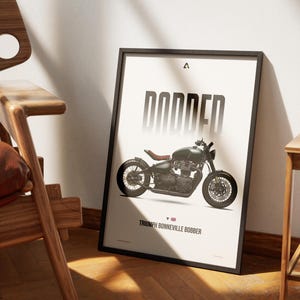 May include: Framed poster featuring a Triumph Bonneville Bobber motorcycle. The poster has a black frame and displays the word "BOBBED" in large, bold letters above the motorcycle image. The motorcycle is olive green with a brown seat. The text "Triumph Bonneville Bobber" is at the bottom.