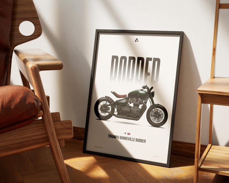 May include: Framed poster featuring a Triumph Bonneville Bobber motorcycle. The poster has a black frame and displays the word "BOBBED" in large, bold letters above the motorcycle image. The motorcycle is olive green with a brown seat. The text "Triumph Bonneville Bobber" is at the bottom.