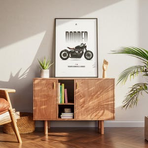 May include: A framed poster with a black and white motorcycle illustration, labeled "DODDER" and "Triumph Bonneville Bobber." The poster is displayed on a wooden cabinet with a plant, books, and decorative items. The room has a modern aesthetic.
