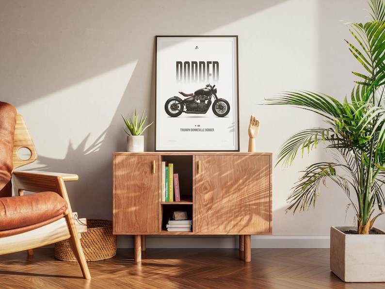May include: A framed poster with a black and white motorcycle illustration, labeled "DODDER" and "Triumph Bonneville Bobber." The poster is displayed on a wooden cabinet with a plant, books, and decorative items. The room has a modern aesthetic.